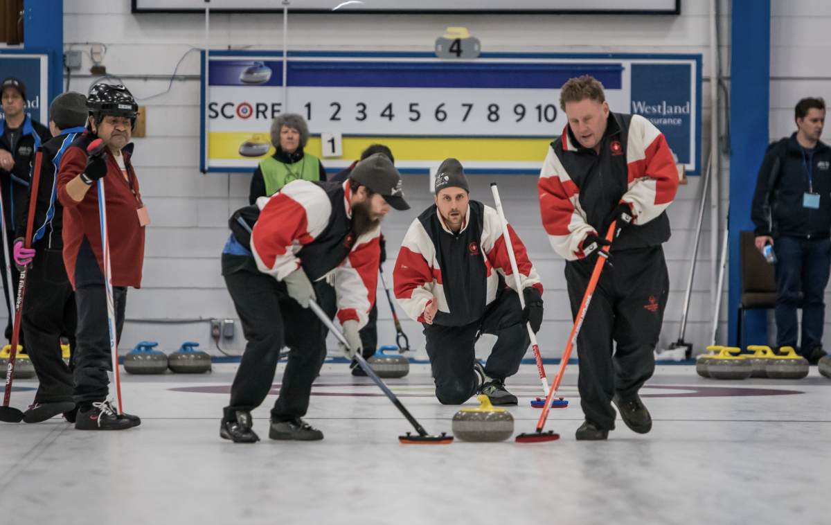 Curling action at an indoor ice rink. A group of players, some wearing red and white jackets, are focused on guiding a yellow curling stone across the ice. One man in the foreground is crouched low, intensely watching the stone's path, while two others sweep with brooms to influence the stone's trajectory. In the background, there's a scoreboard and other participants observing the game. Advertisements for 'Westland Insurance' can be seen on the rink wall.